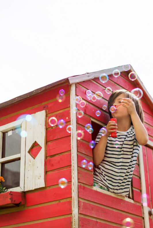 Lapsi puhaltaa saippuakuplia punaisen leikkimökin ikkunasta